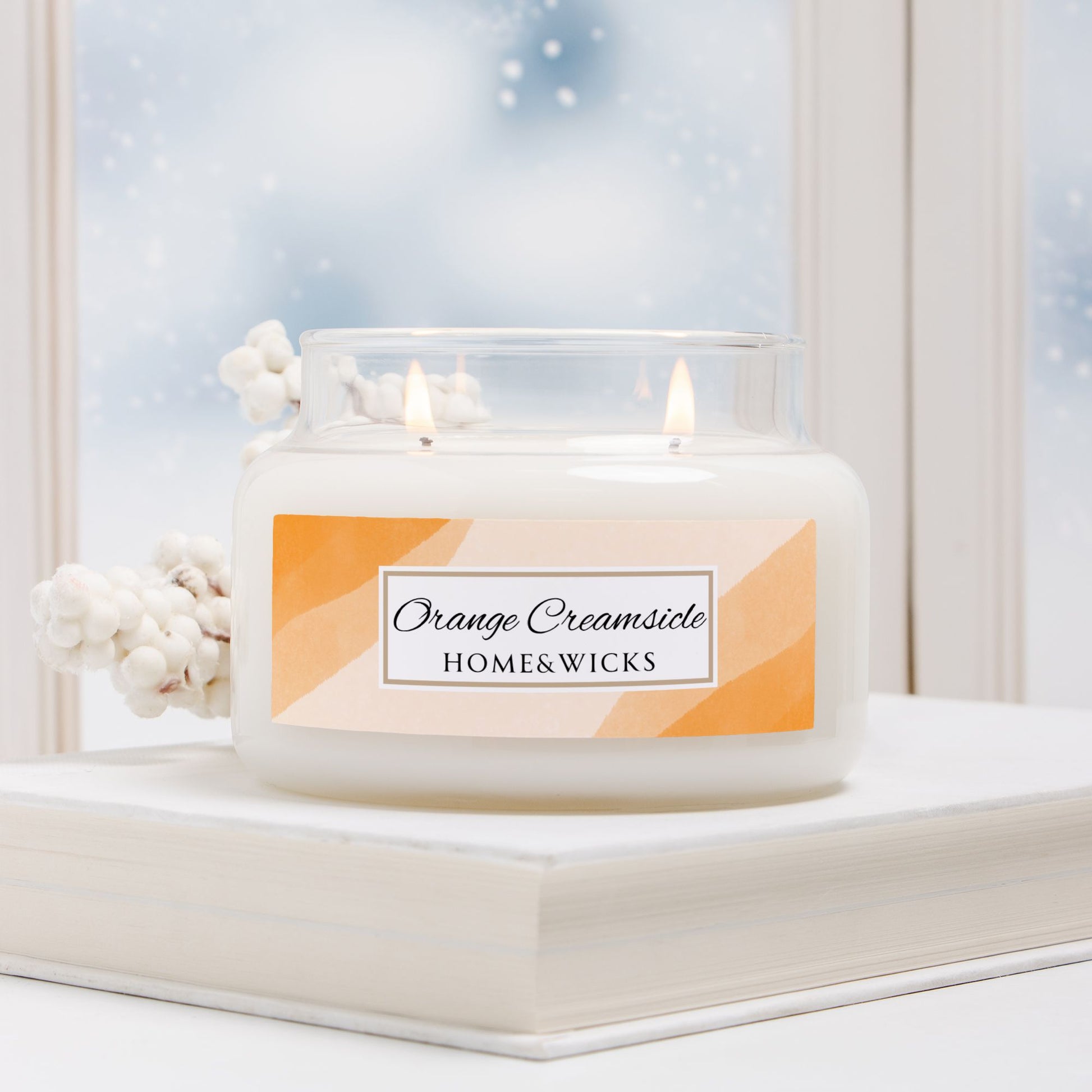 Candle labeled 'Orange Creamsicle' by HOME&WICKS on a windowsill with snow outside.