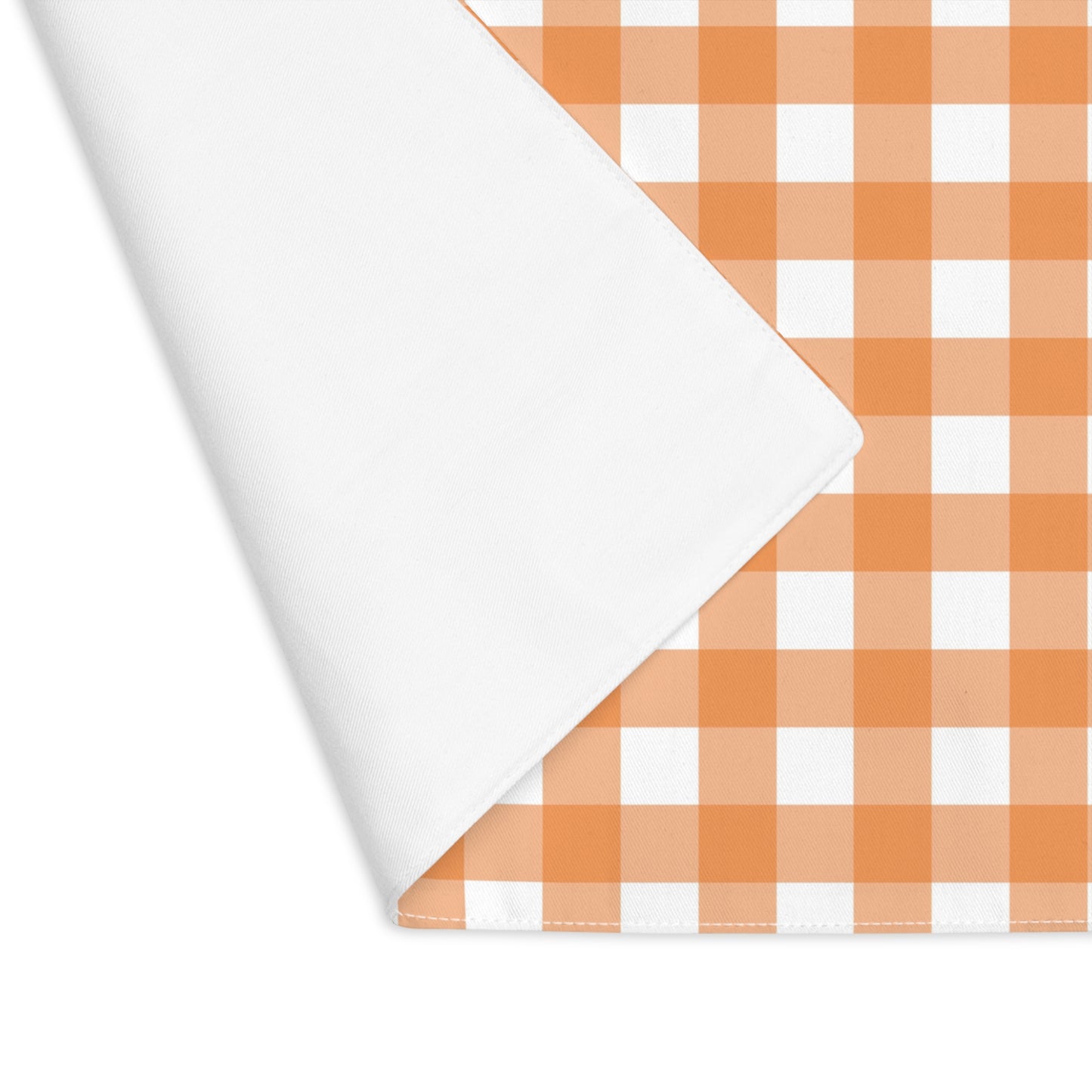 The corner is folded over to reveal white back of our Orange Gingham placemat with an orange and white checkered pattern on the table runner fabric on a white backaground
