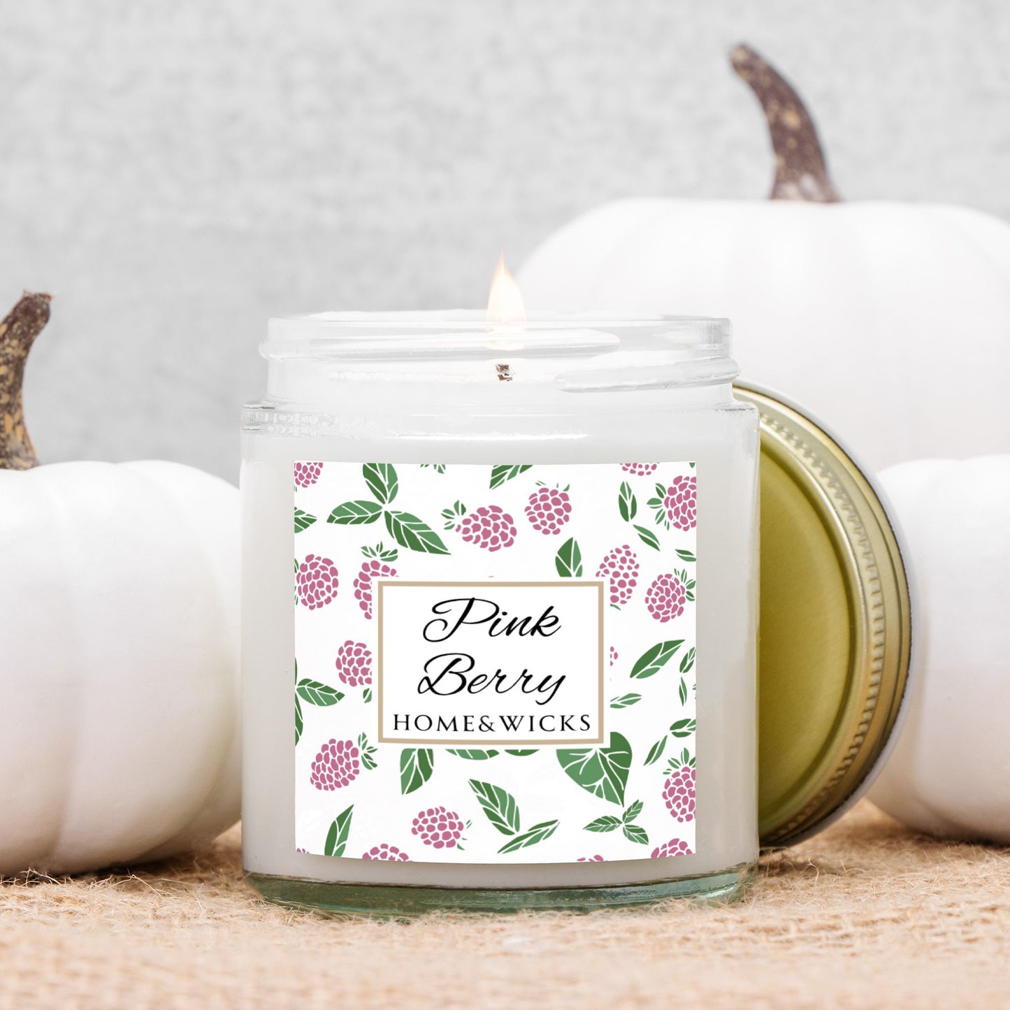 Homes & Wicks Pink Berry candle in a clear jar with gold lid  with pink berry label graphics on a white background, surrounded by white pumpkins.