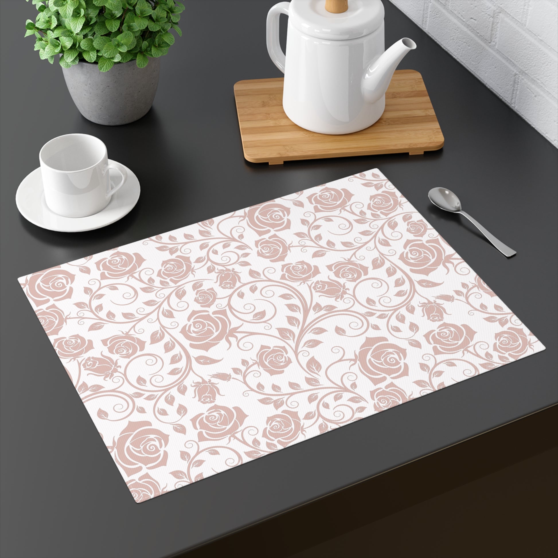 Floral pink rose patterned placemat on a dark surface with a teapot, cup, and spoon.