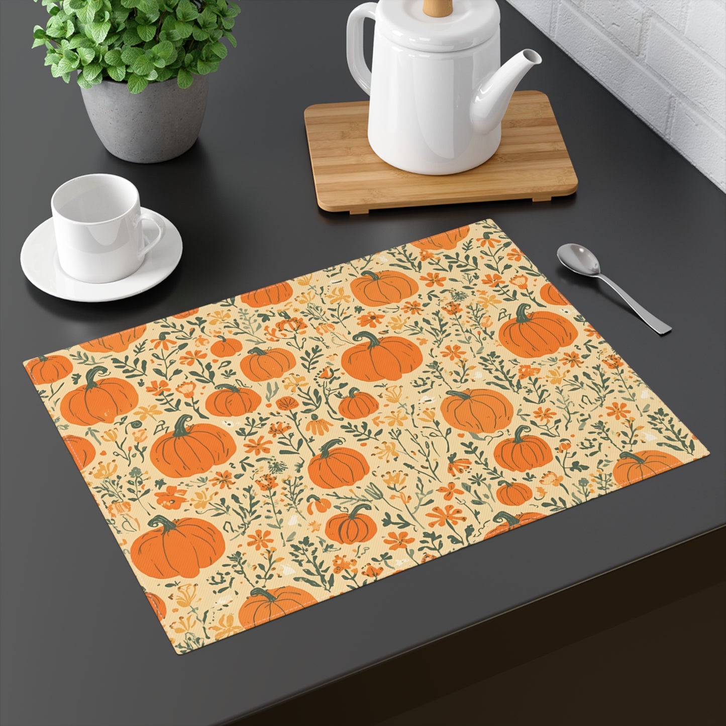 Our Pumpkin Garden cotton twill placemat has an orange pumpkins and flowers placemat design on a beige background on a dark surface with tea set and plant.