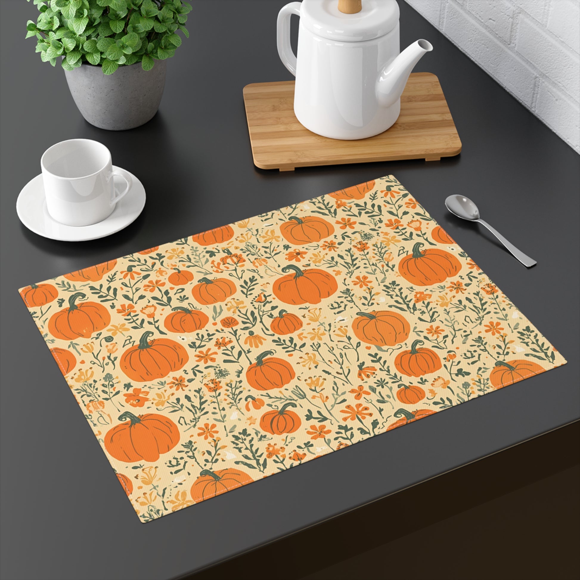 Our Pumpkin Garden cotton twill placemat has an orange pumpkins and flowers placemat design on a beige background on a dark surface with tea set and plant.