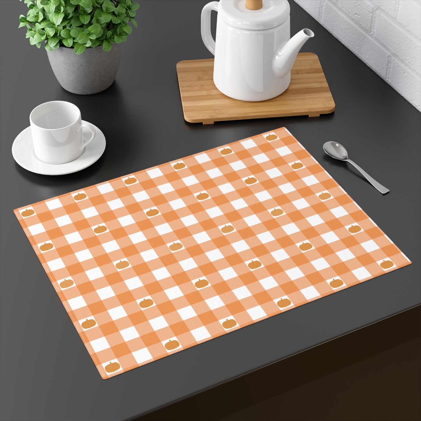 Our Pumpkin Gingham placemat has an orange and white checkered pattern with small pumpkins on the cotton twill placemat fabric on a dark surface with a teapot, cup, and spoon.