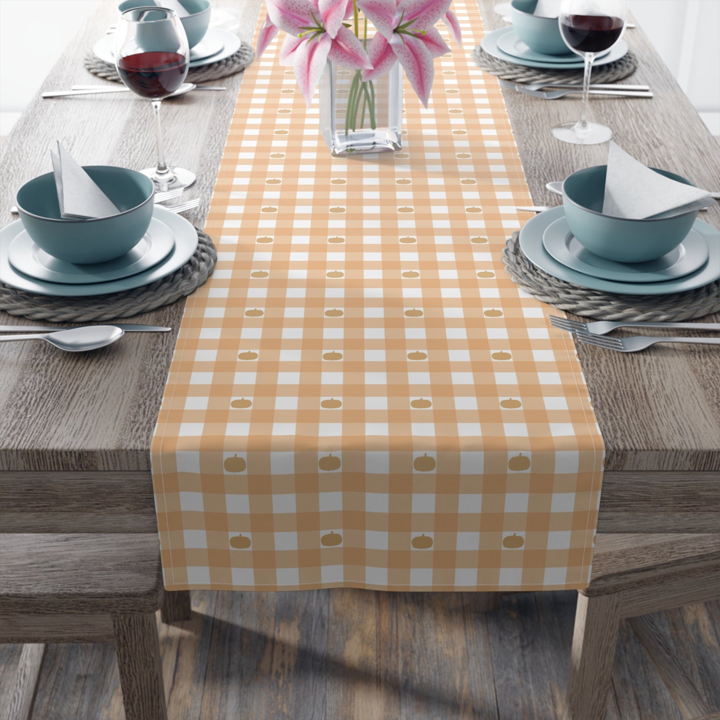 Our cotton twill Pumpkin Gingham table runner has an orange and white checkered pattern with small pumpkins on the table runner fabric  and sits on a wooden table with place settings and flowers.