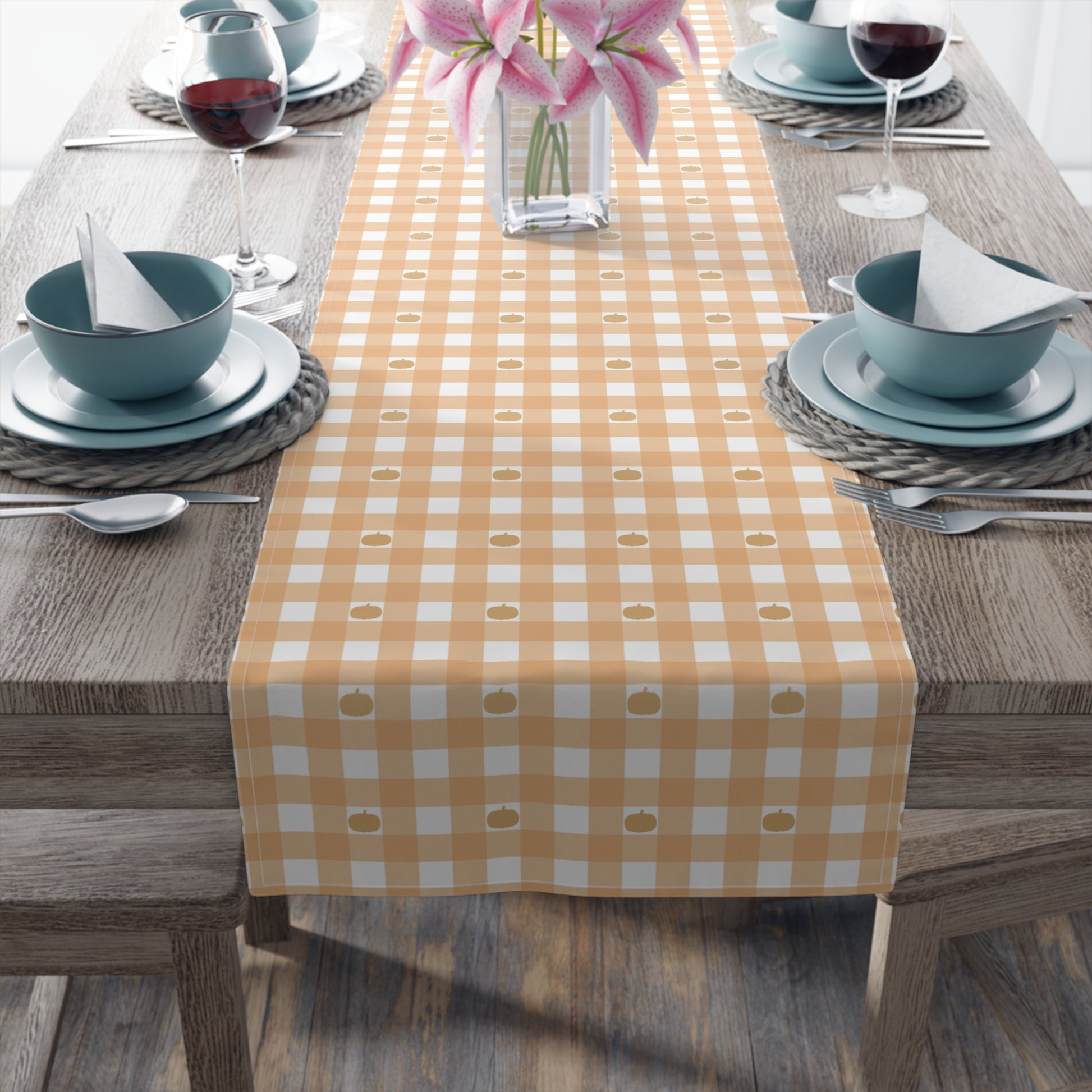 Our cotton twill Pumpkin Gingham table runner has an orange and white checkered pattern with small pumpkins on the table runner fabric  and sits on a wooden table with place settings and flowers.