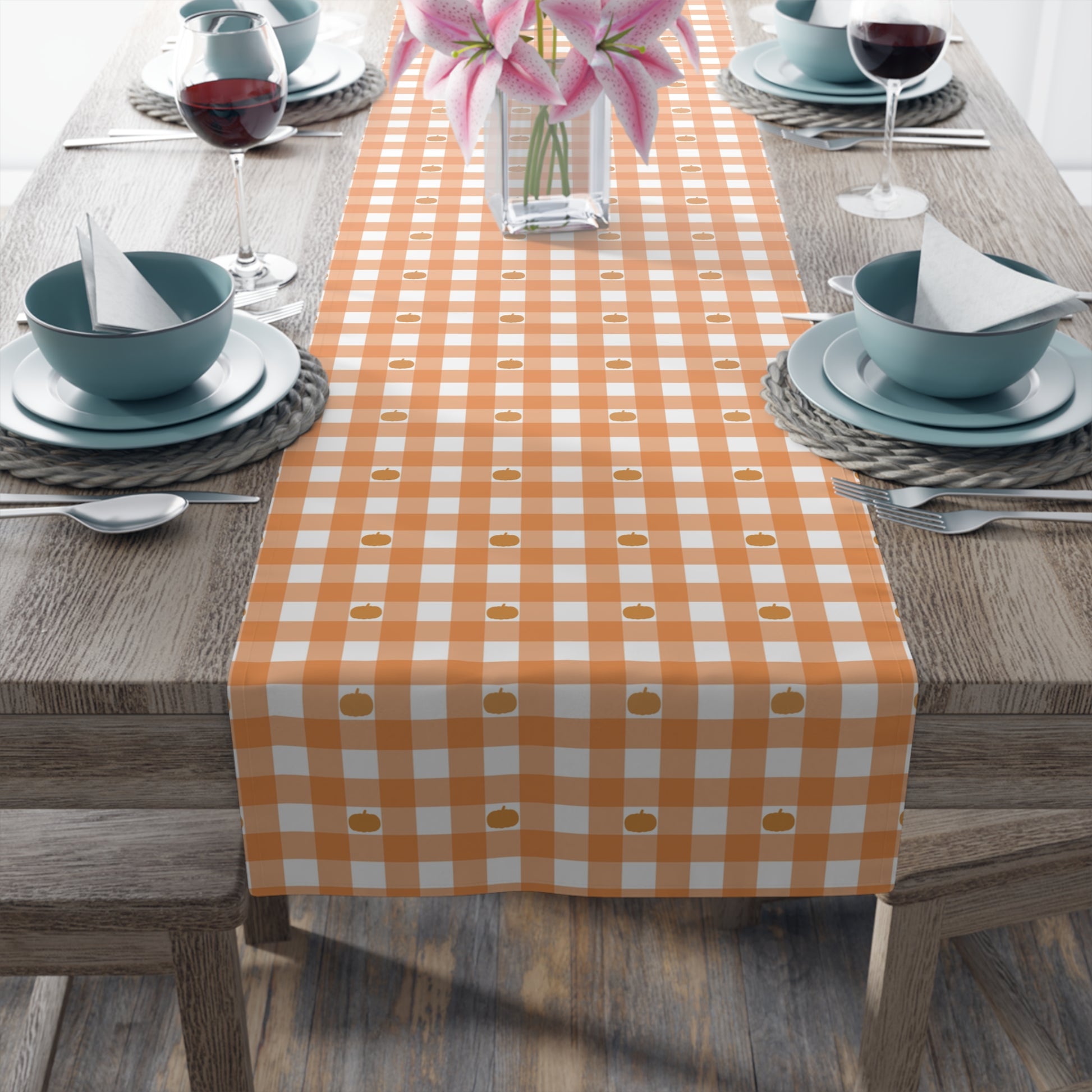 Our Pumpkin Gingham table runner has an orange and white checkered pattern with small pumpkins on the table runner fabric and sits on a table setting with an orange and white checkered table runner on a wooden table.