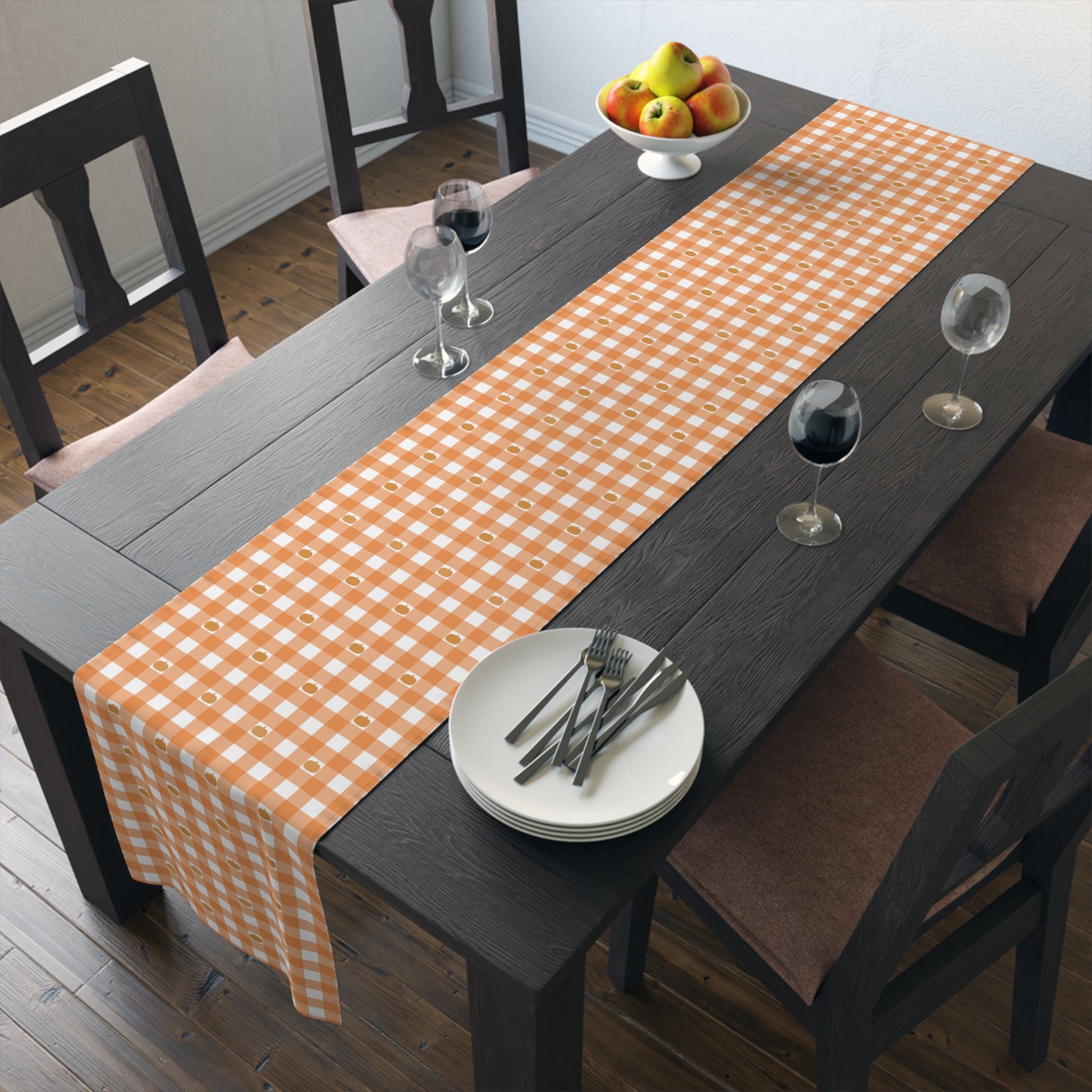 Our polyester Pumpkin Gingham table runner has an orange and white checkered pattern with small pumpkins on the table runner fabric and sits on a dining table with plates, and glasses.