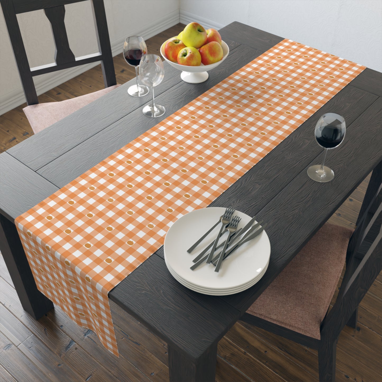 Our polyester Pumpkin Gingham table runner has an orange and white checkered pattern with small pumpkins on the table runner fabric and sits on a dark brown wood table with plates, forks, and a fruit bowl.