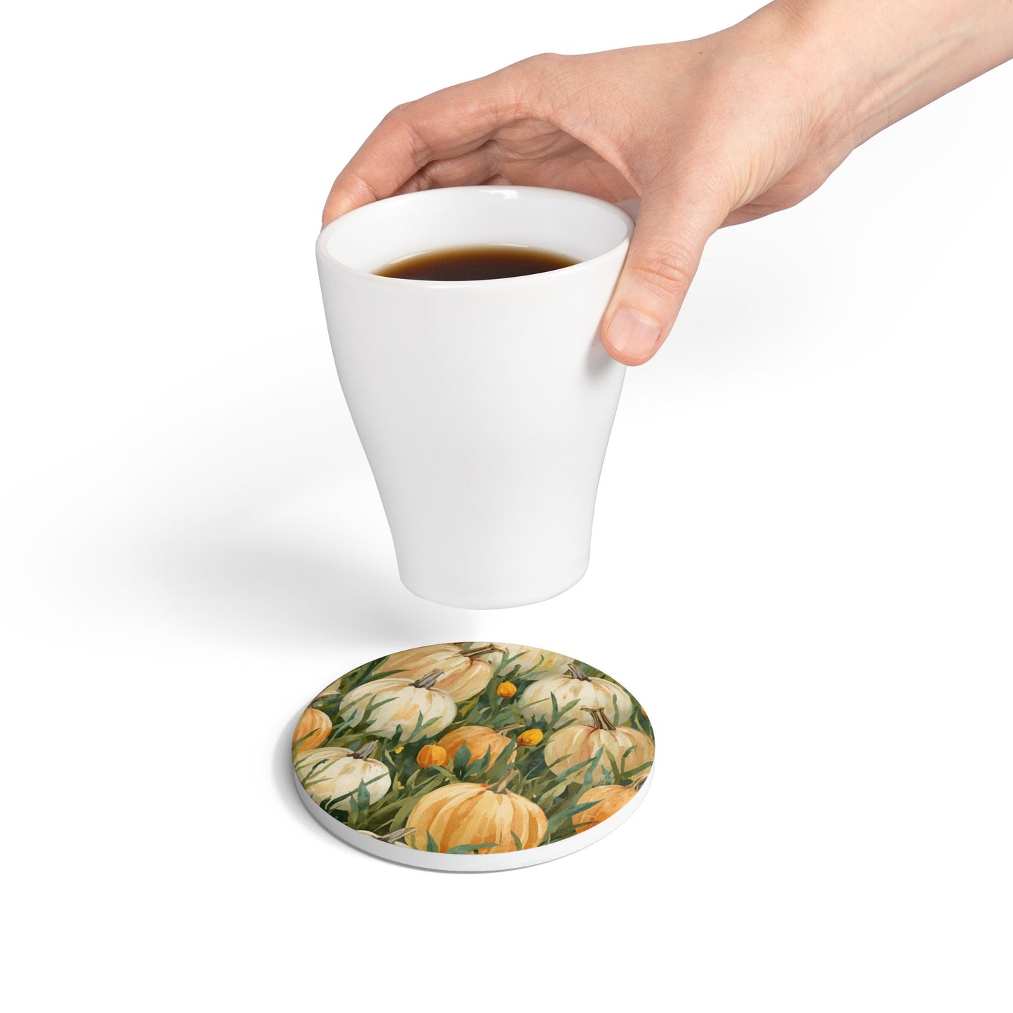 Pumpkin Patch round ceramic tile coaster has white and orange pumpkins in grass with orange tulips printed on top of the tile coaster and a hand holding a white mug above coaster on a white background