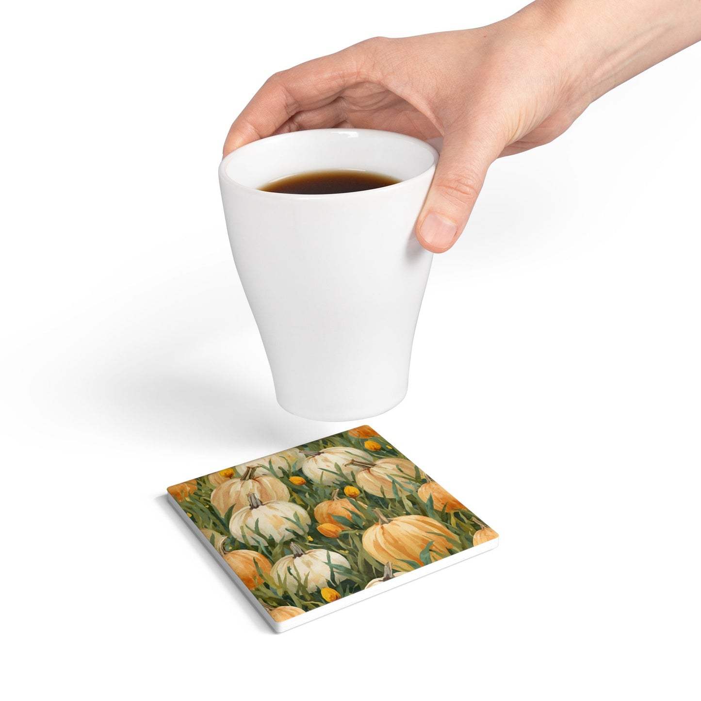 Pumpkin Patch ceramic tile coaster has white and orange pumpkins in grass with orange tulips printed on top of the tile coaster and a hand above coaster holds a coffee cup on a white background