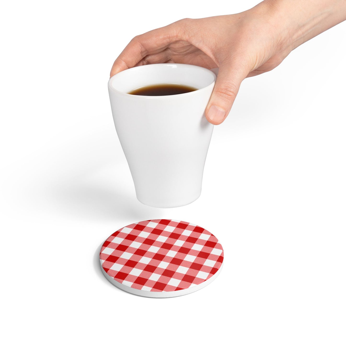Red gingham round ceramic tile coaster with red and white checkered pattern on top of coaster, with hand holding coffee cup above coaster, on a white background.