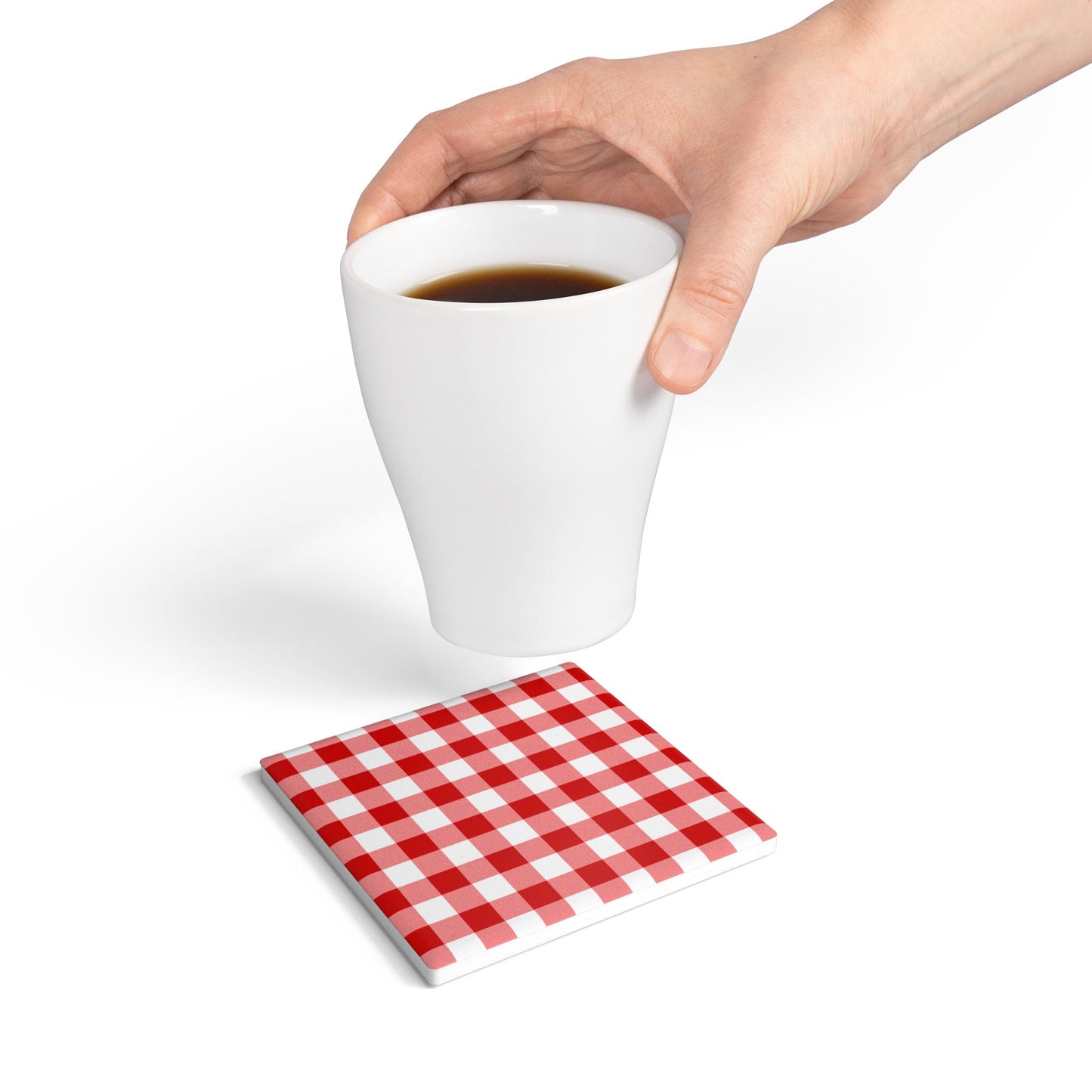 Red gingham square ceramic tile coaster with red and white checkered pattern on top of coaster, with hand holding coffee cup above coaster, on a white background.
