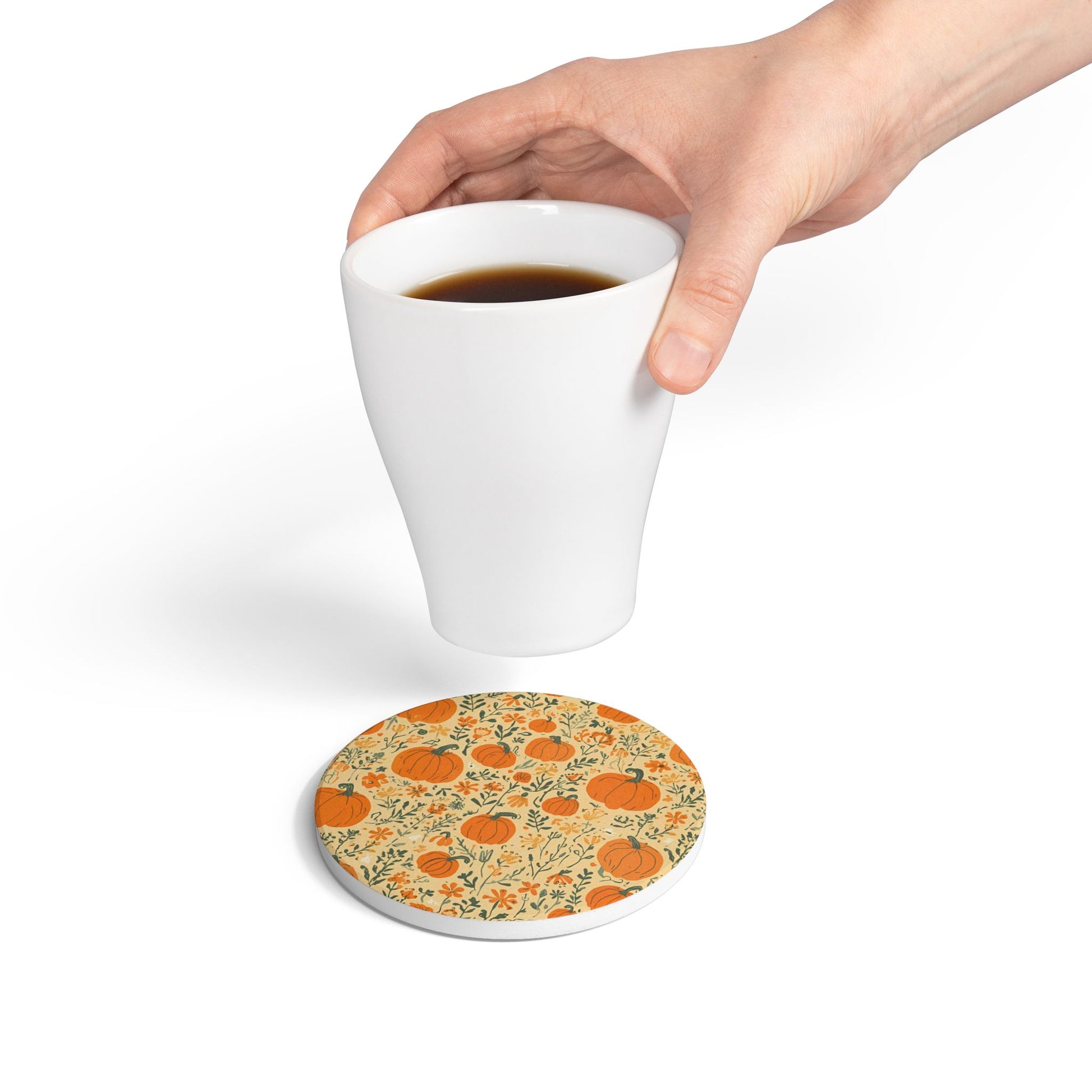 Hand holding a white mug above a pumpkin and floral patterned ceramic tile coaster on a white background
