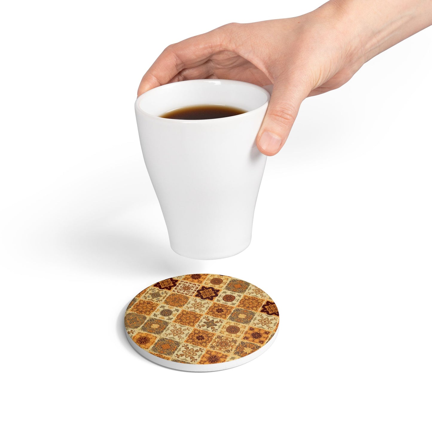 Hand holding a white mug with a brown, gold and grayish-green floral mosaic patterned round tile coaster underneath on a white background