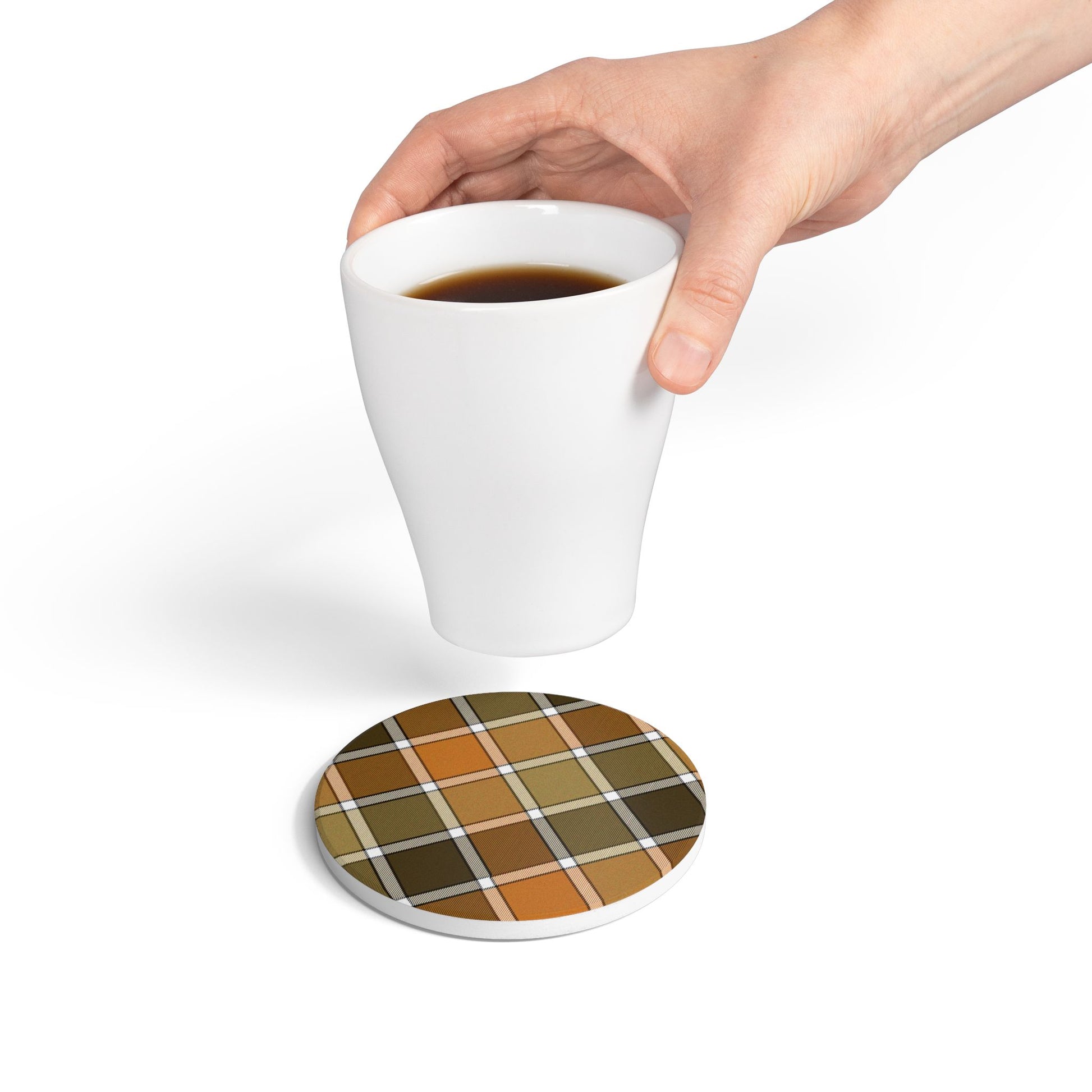Hand holding a white mug with a fall colored plaid round ceramic tile coaster underneath on a white background