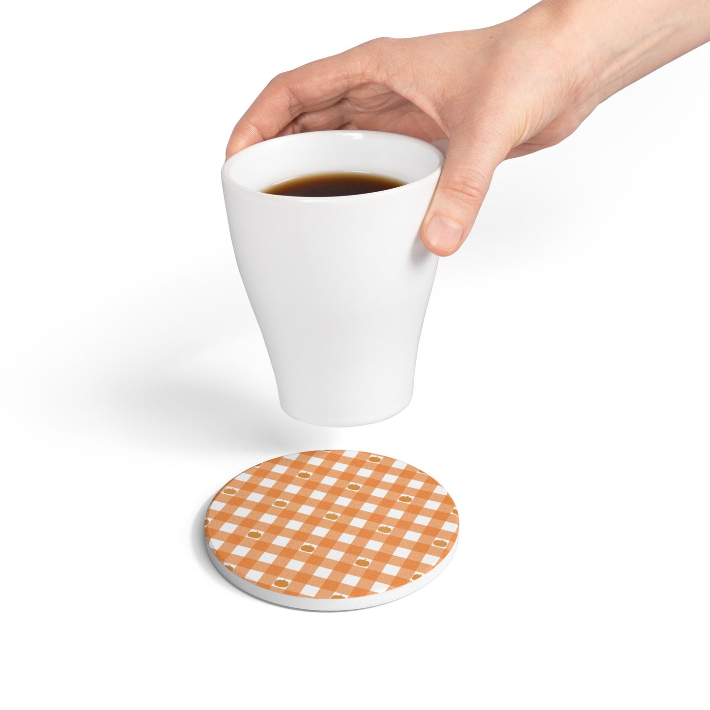 Hand holding a white mug over a round checkered orange gingham with pumpkins ceramic tile coaster on a white background