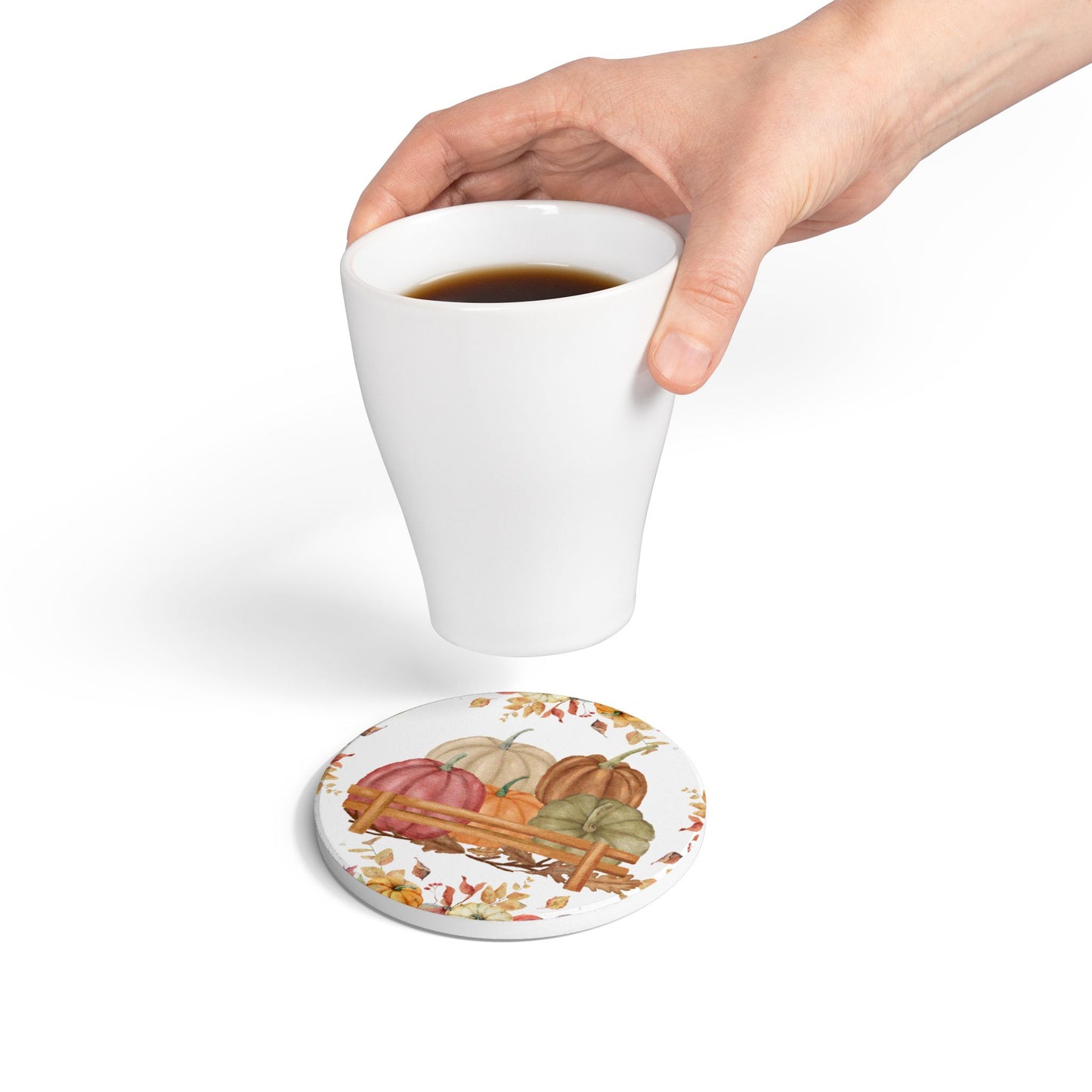 Hand holding a white mug with a colorful round tile coaster featuring pumpkins, gourds, and corn.