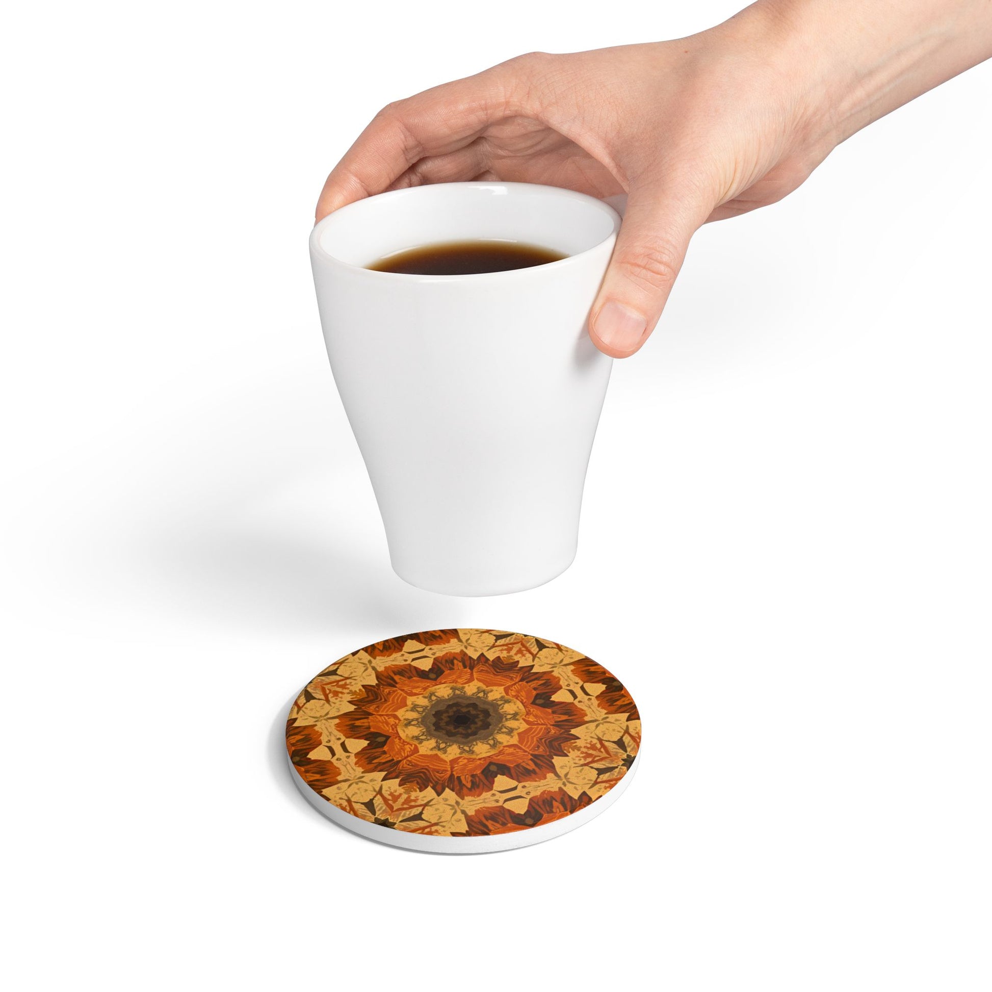 Hand holding a white mug with an orange, gray and yellow mandala design tile coaster underneath on a white background
