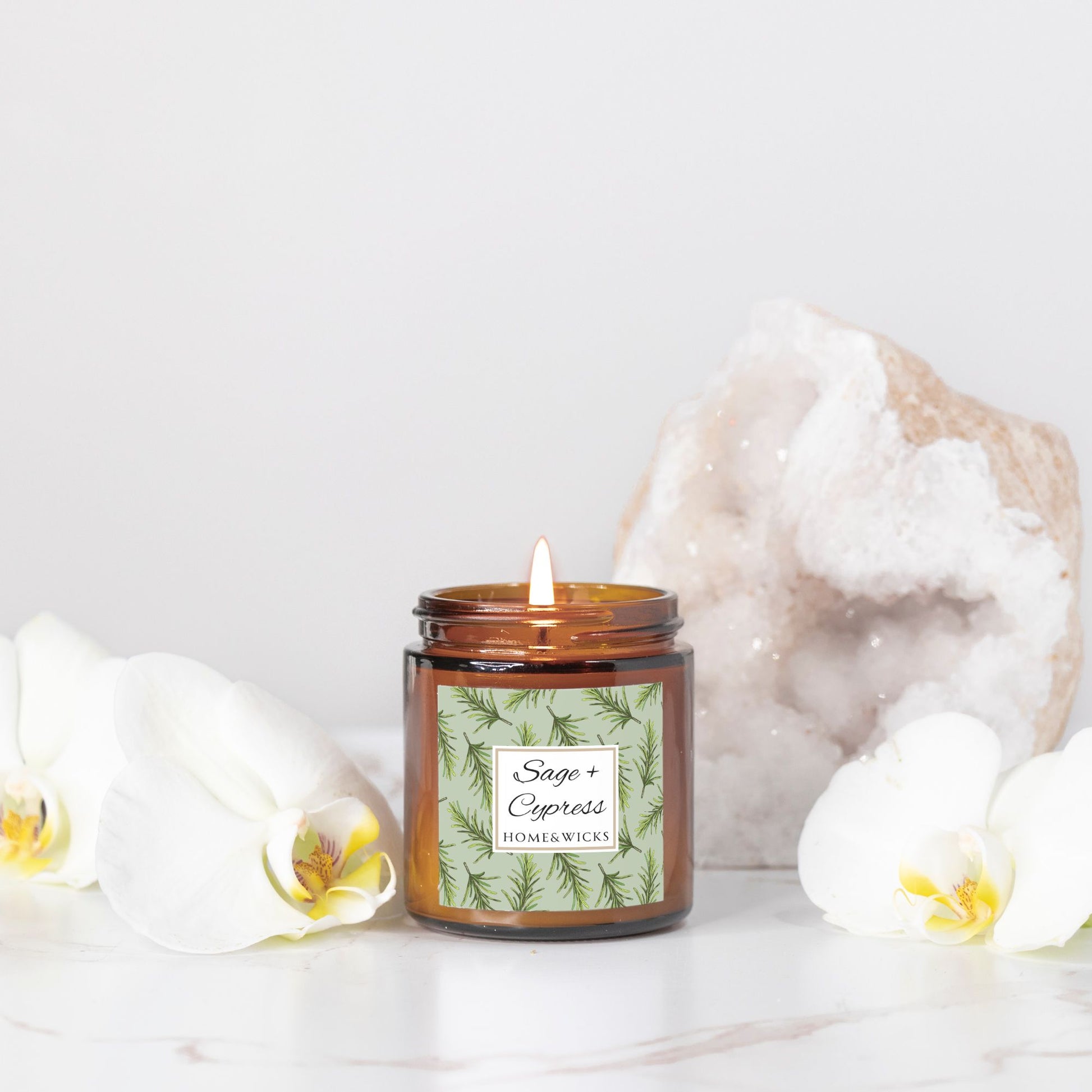 Sage & Cypress candle in a 4 oz. amber glass jar with a gold lid and 'Sage + Cypress' label on a white background by Home & Wicks in front of white flowers and a crystal on a light background