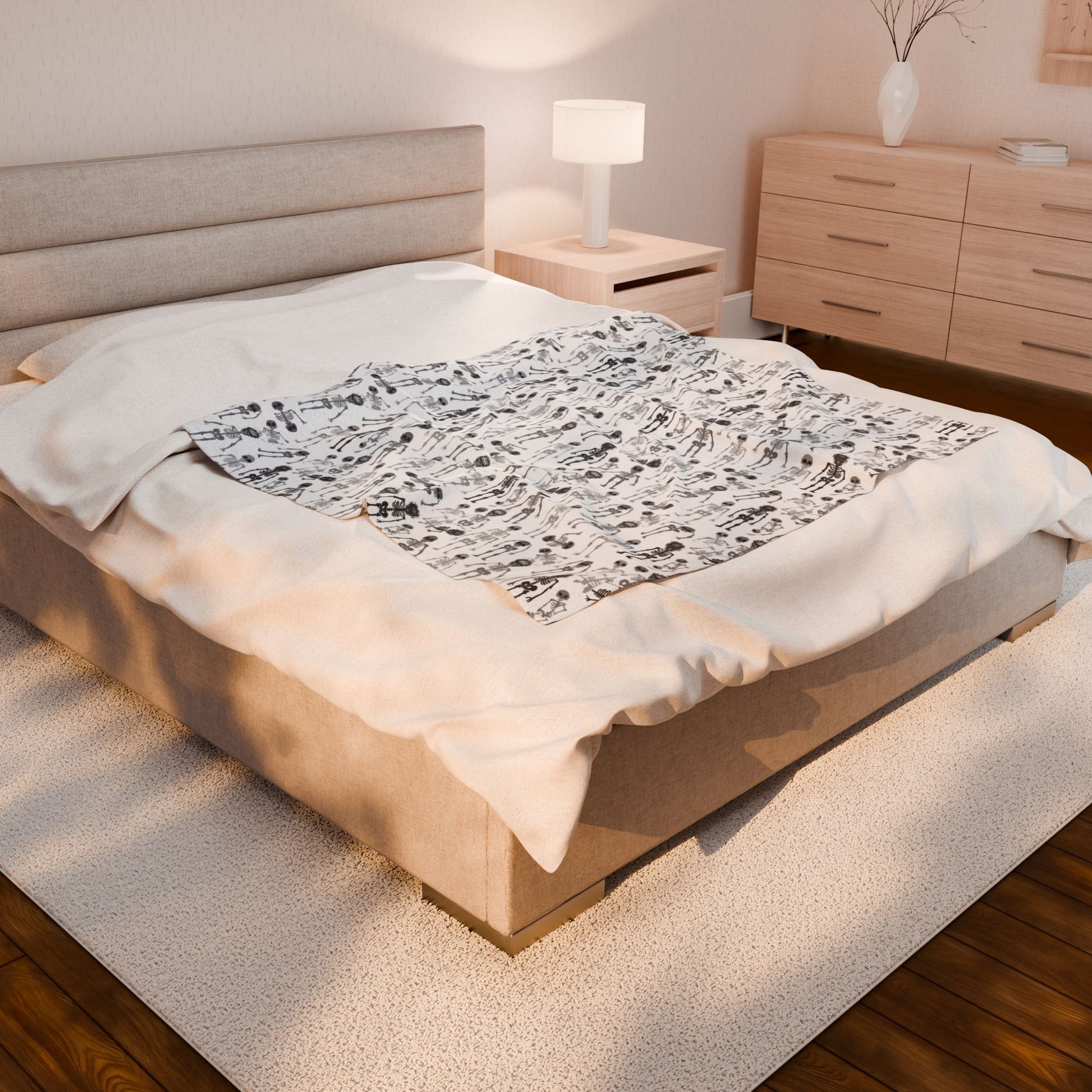 Halloween-themed Skeletons patterned blanket with black and white skeletons on a white background on front side of blanket, laying on a bed in a bedroom with a neatly made bed and wooden furniture.