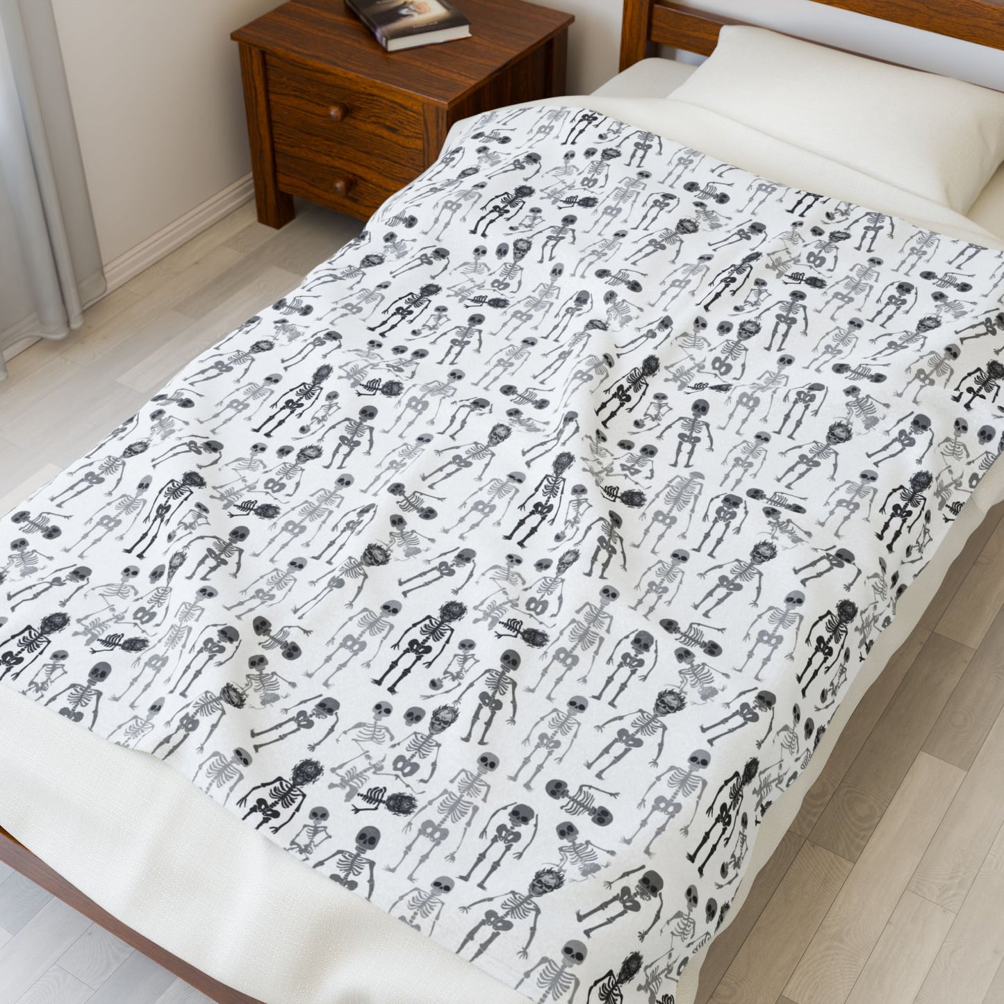Halloween-themed Skeletons patterned blanket with black and white skeletons on a white background on front of blanket, on a bed in a bedroom setting