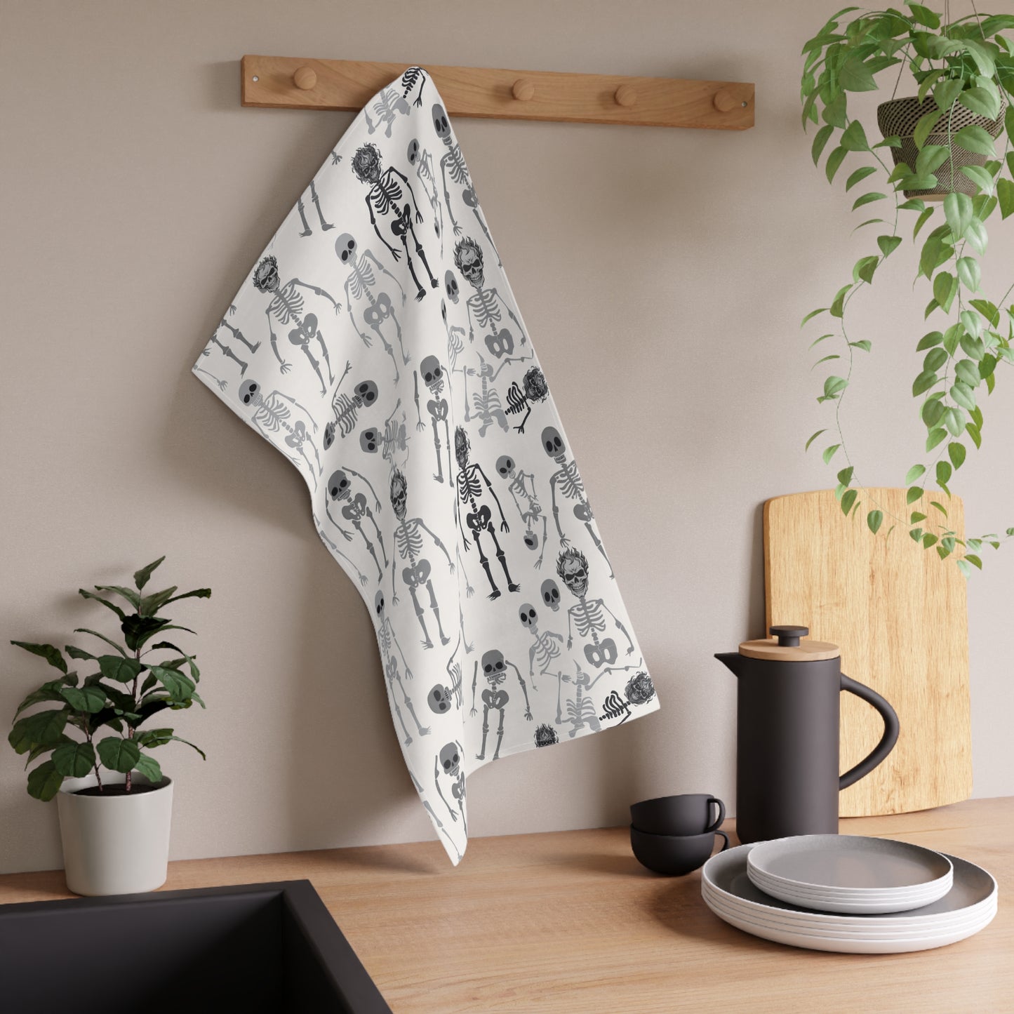 Skeletons tea kitchen towel with black and white skeleton pattern on front of towel, hanging on a rack, next to a plant and kitchen items.