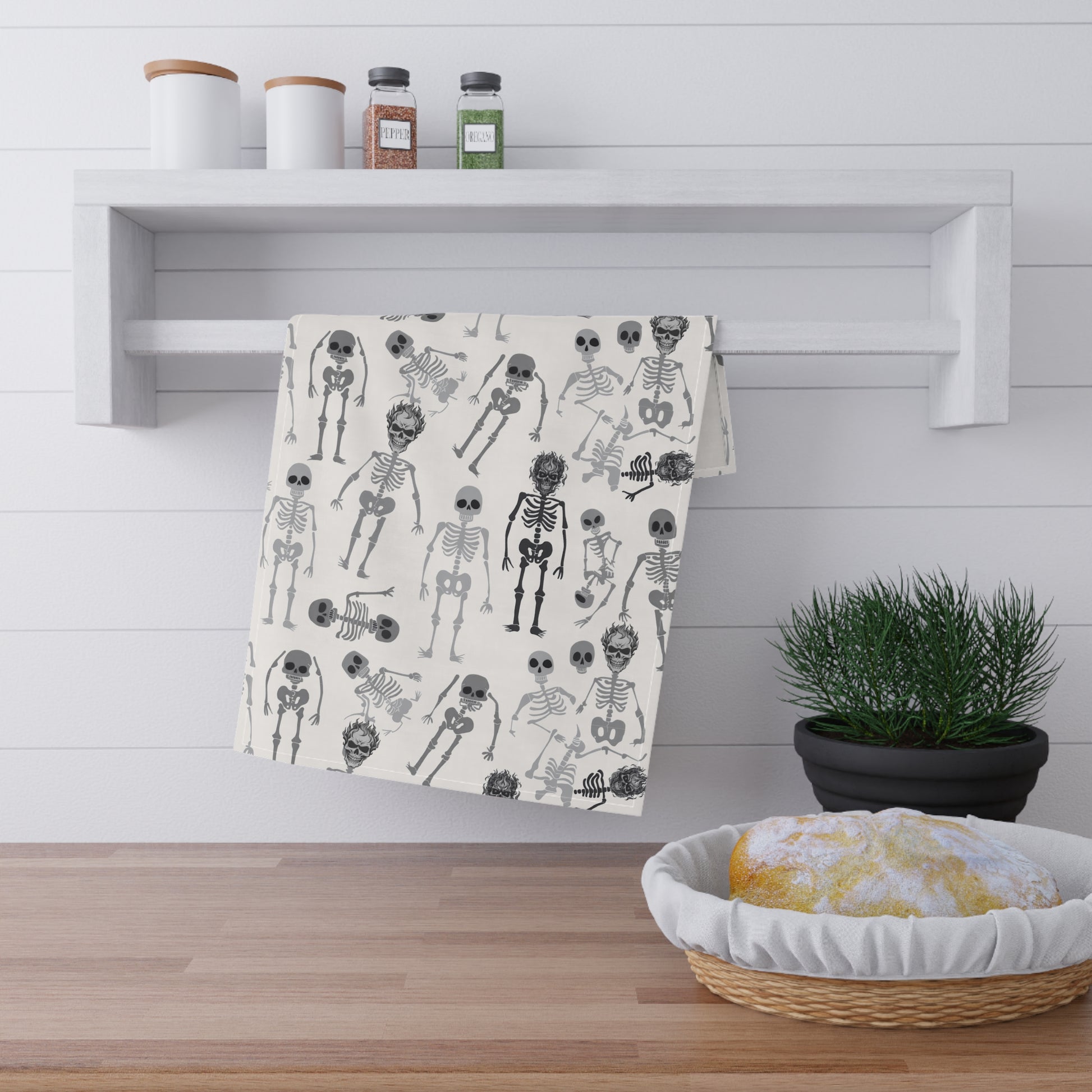 Skeletons tea kitchen towel with black and white skeleton pattern on front of towel, hanging on a wooden shelf with a plant and basket below.