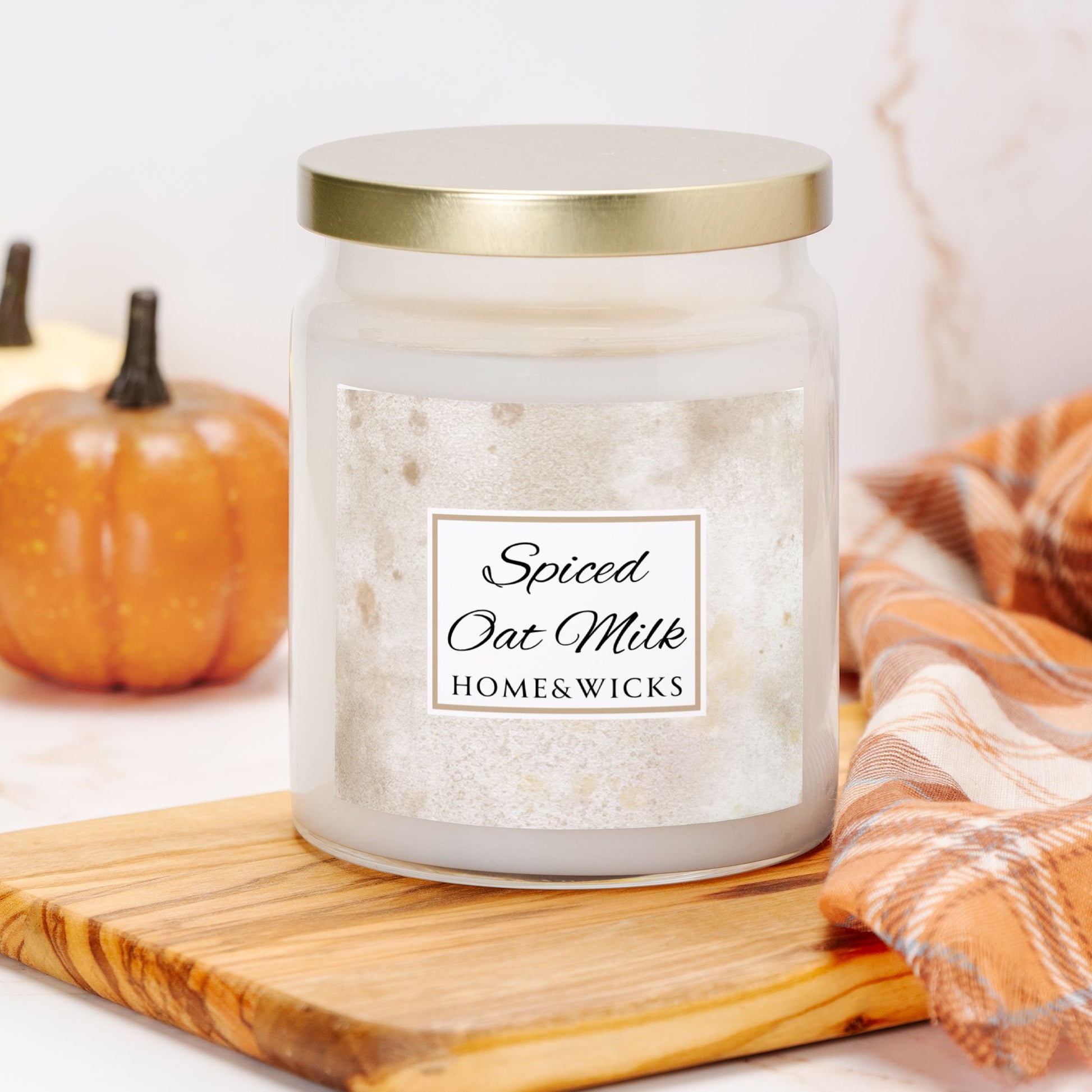 Candle jar labeled 'Spiced Oat Milk' by Home & Wicks on a wooden board with pumpkins and a plaid cloth in the background.
