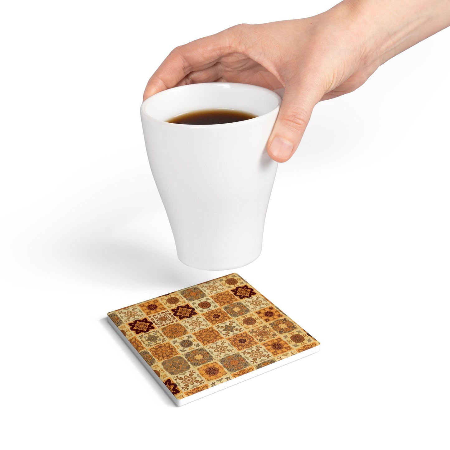 Hand holding a white mug with a square ceramic tile coaster with fall flower mosaic pattern underneath on a white background