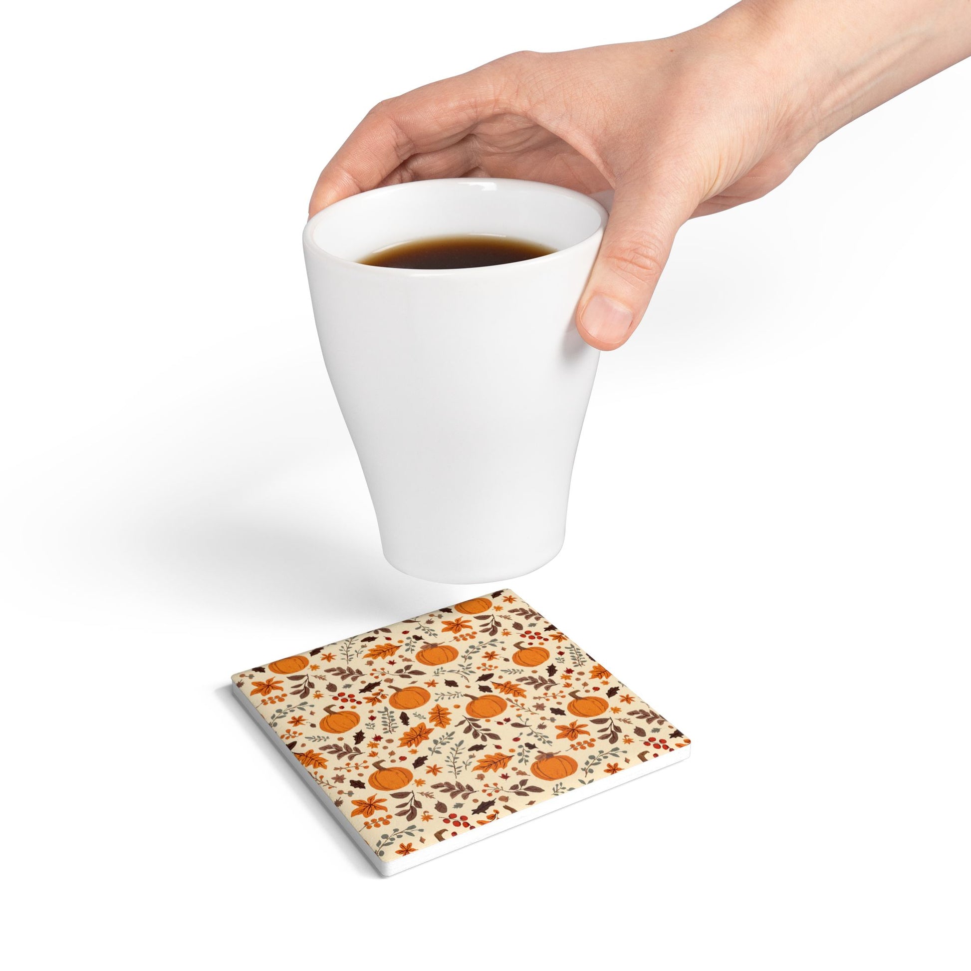 Hand holding a white mug with a square ceramic tile coaster underneath with fall leaves and pumpkins design on a white background