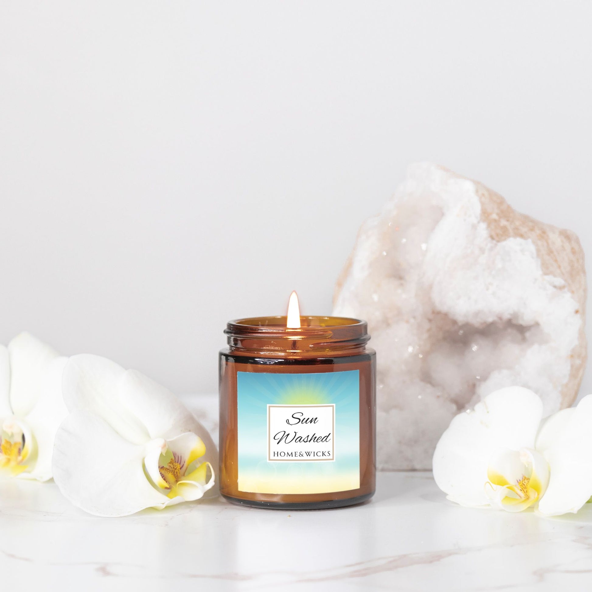 Sun Washed candle in a 4 oz.  jar labeled 'Sun Washed' with sun graphic by Home & Wicks on a white background, surrounded by white flowers and a crystal on a light background