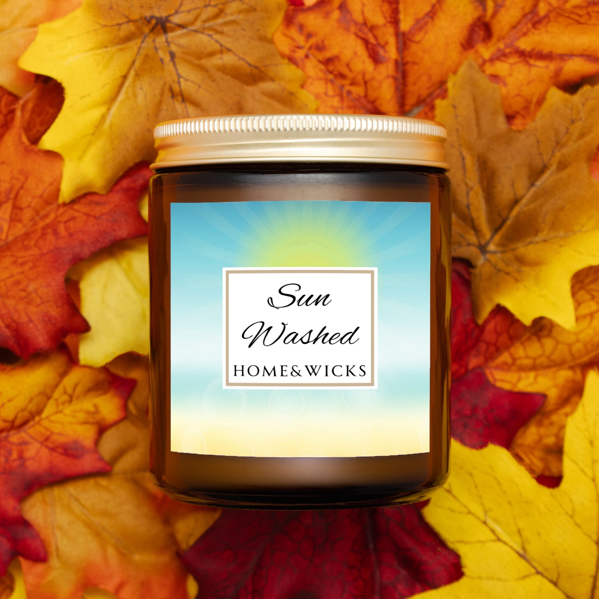 Candle in a 9 oz. amber jar with gold lid with 'Sun Washed' label on with sun graphic gradient background by Home & Wicks on a bed of autumn leaves.