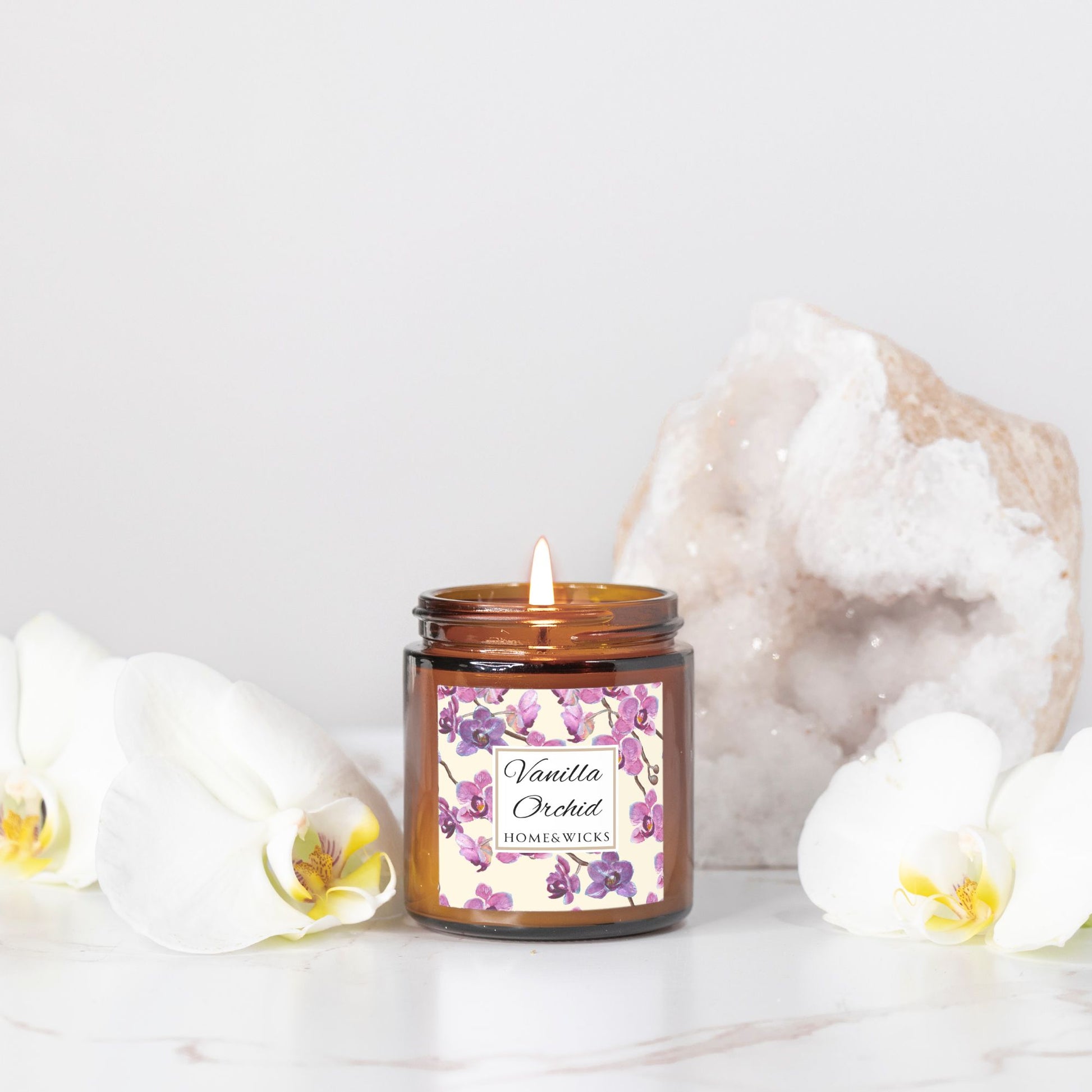 Vanilla Orchid candle in a 4 oz. amber glass candle jar with gold lid with a pink and purple orchid floral label on a white background with rock and orchid flower decor by Home & Wicks