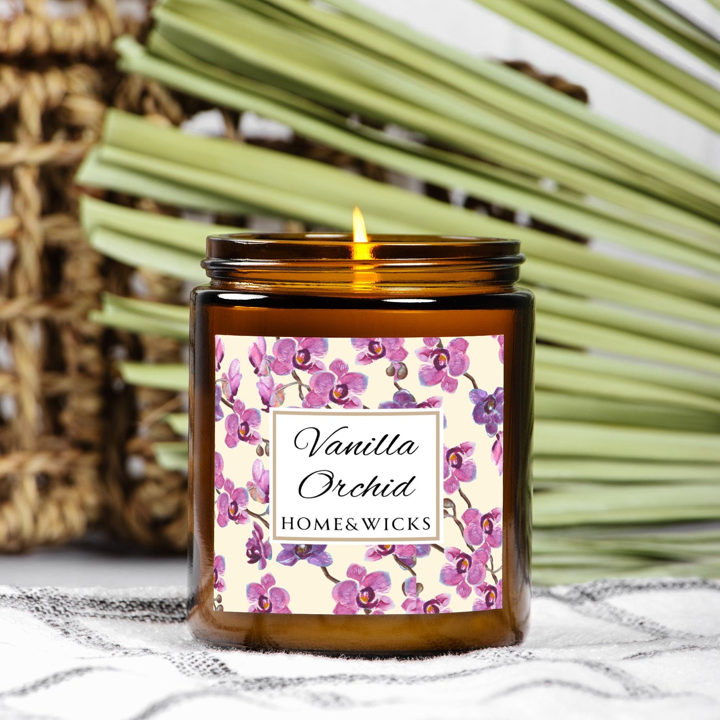Vanilla Orchid candle in an amber jar with gold lid with a pink and purple orchid design label by Home & Wicks with wicker basket and palm leaves in the background