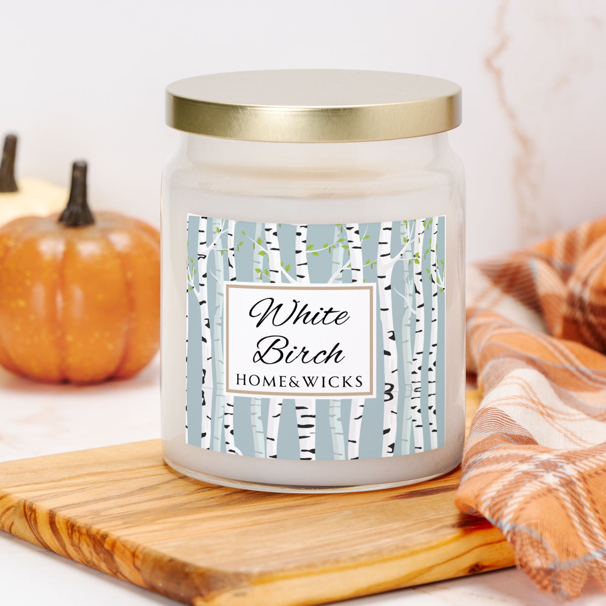 Candle jar labeled 'White Birch' by Home & Wicks on a wooden cutting board with pumpkins and a plaid cloth in the background.