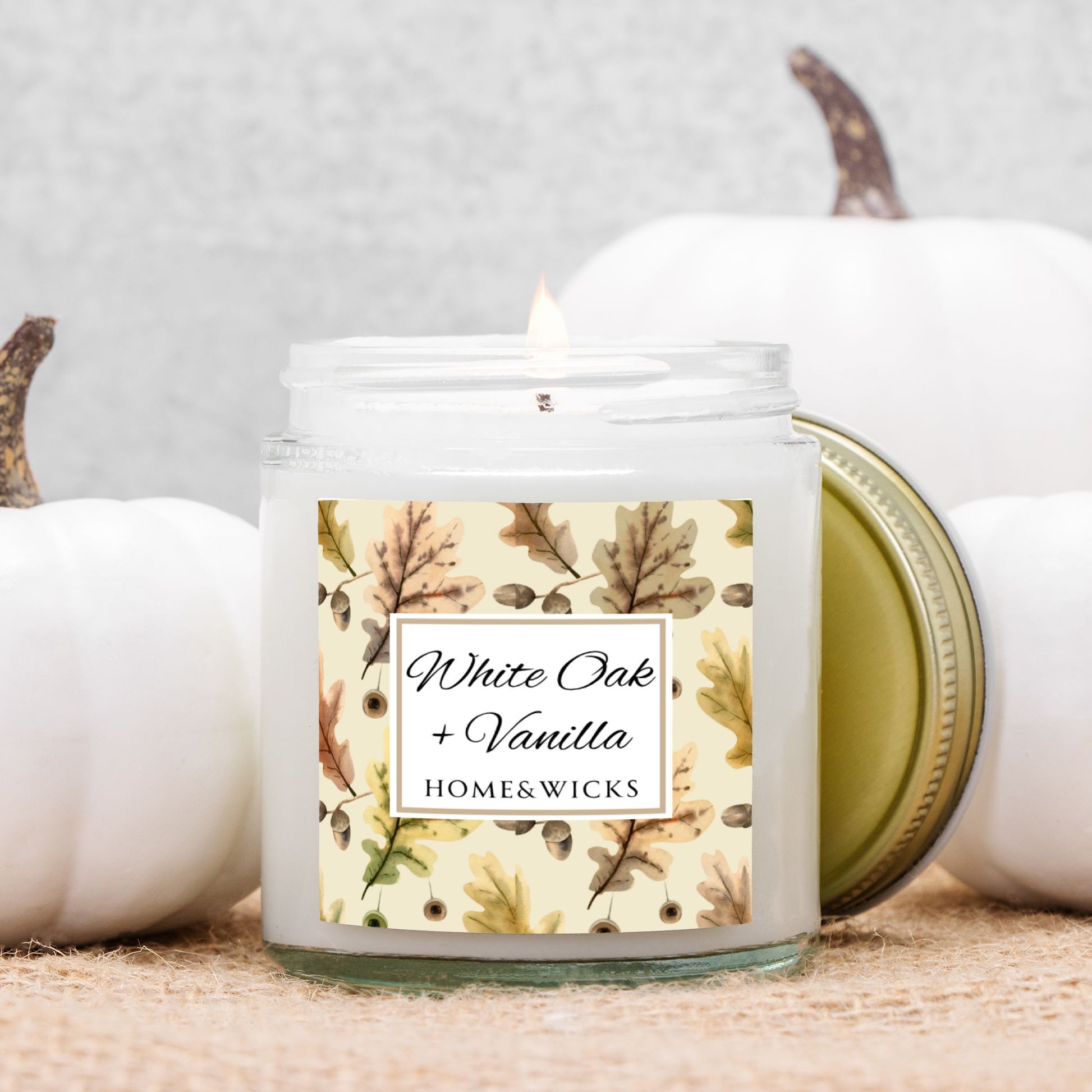 White Oak & Vanilla candle in a 4 oz. clear jar and gold lid with a decorative oak leaf design label featuring leaves and the brand 'Home & Wicks' with pumpkins in the background