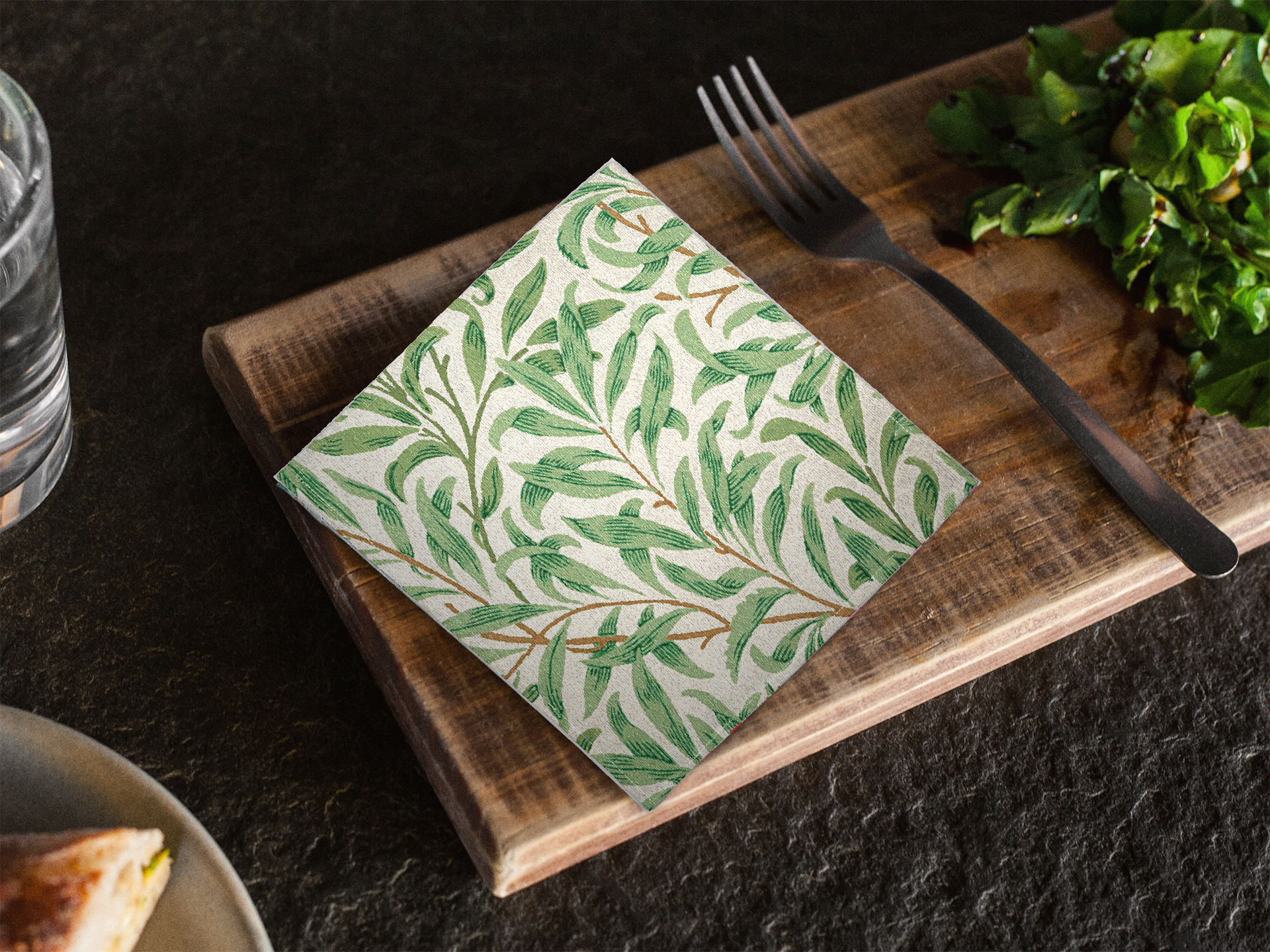 Willow Boughs 10 x 10 cocktail napkin with green leaf willow branch pattern on a wooden cutting board with a fork and water bottle in the background.