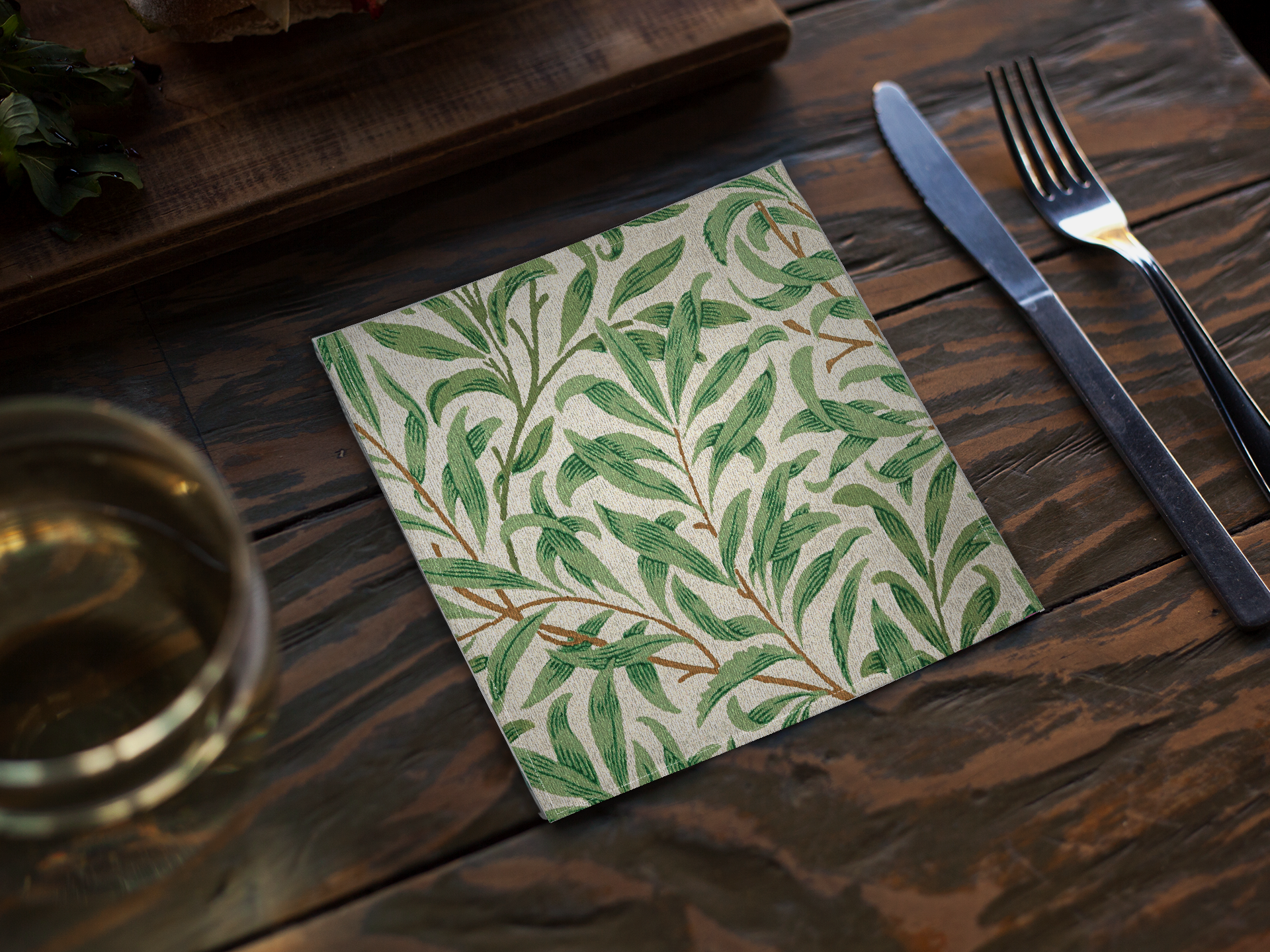 Willow Boughs 10 x 10 cocktail napkin with green leaf willow branch pattern on a wooden table with cutlery and glass.