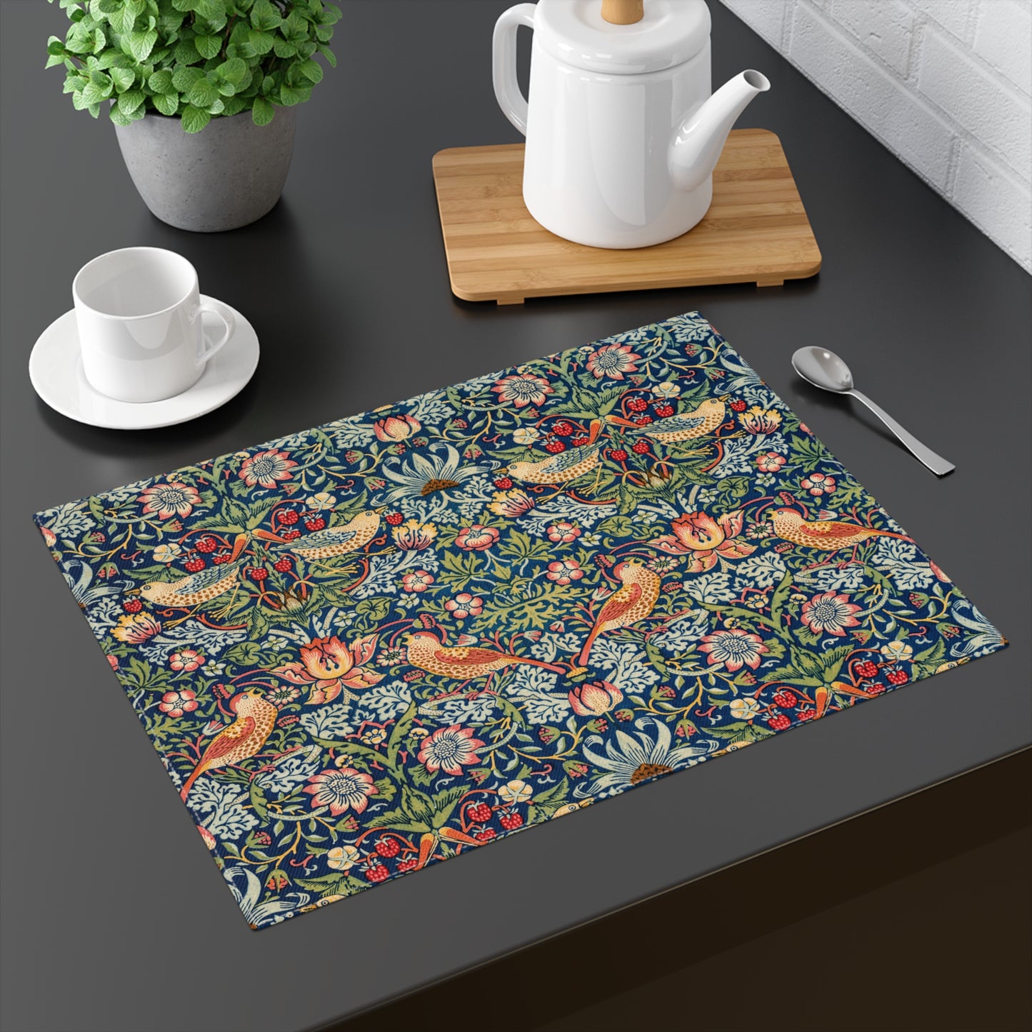 Cotton twill placemat that has the Strawberry Thief illustration by William Morris with a blue background printed on the front of the placemat, sitting on a dark table top, with a teapot and cup in the background.